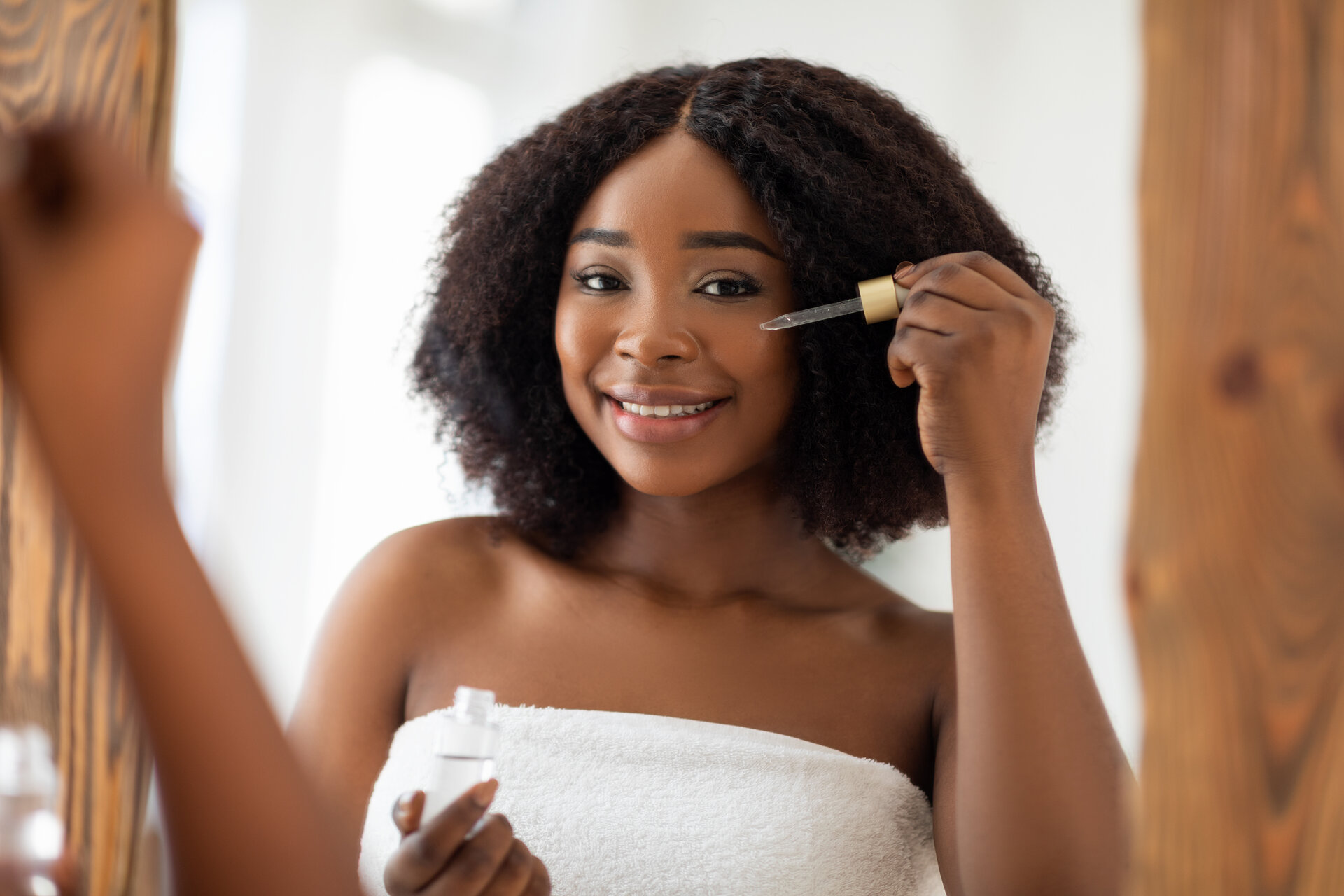 Lächelnde junge schwarze Frau im Handtuch, die ein Gesichtsserum in der Nähe des Spiegels benutzt und sich um ihre Schönheit zu Hause kümmert