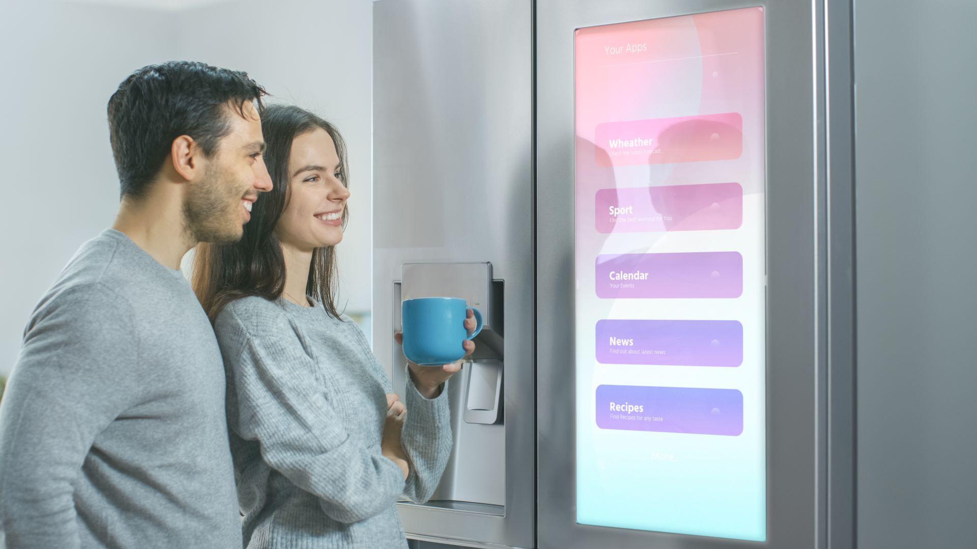 Schönes junges Paar trinkt morgens Kaffee in der Küche. Sie überprüfen die Wettervorhersage und eine Aufgabenliste auf einem intelligenten Kühlschrank zu Hause. Die Wohnung ist hell und gemütlich.